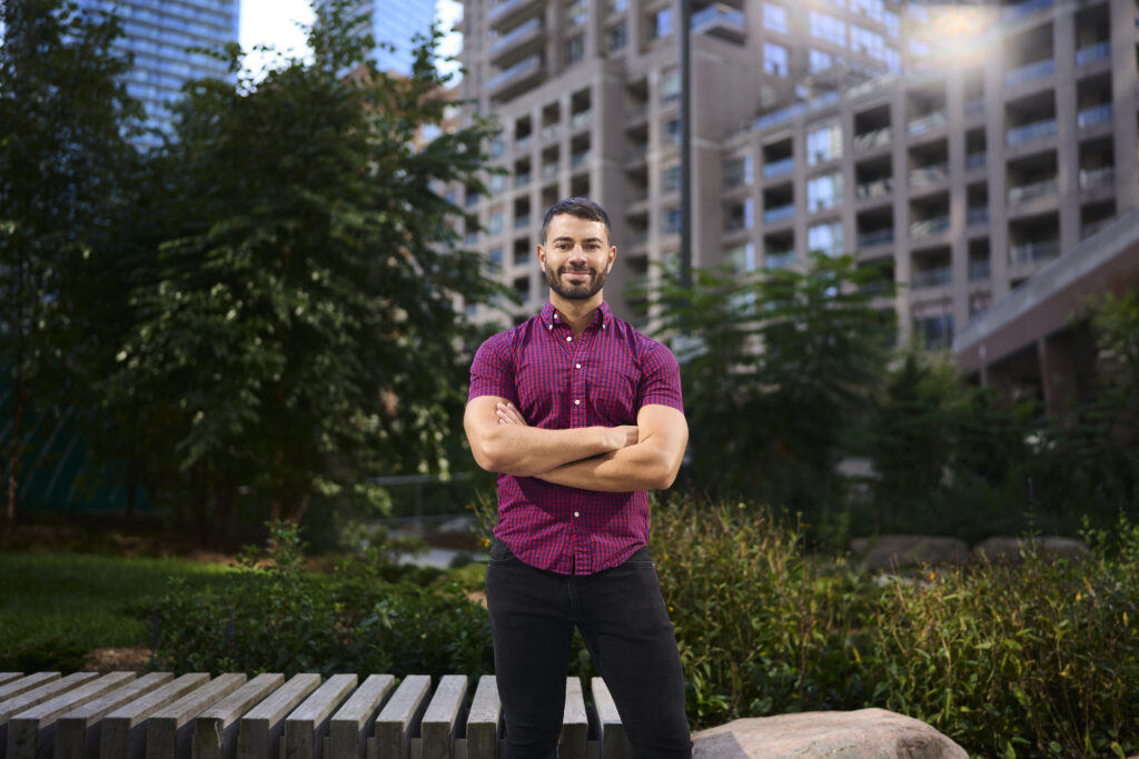 Michael stands confidently with arms crossed wearing a red plaid shirt and black pants. He's standing in a park surrounded by deep greenery and tall city buildings.